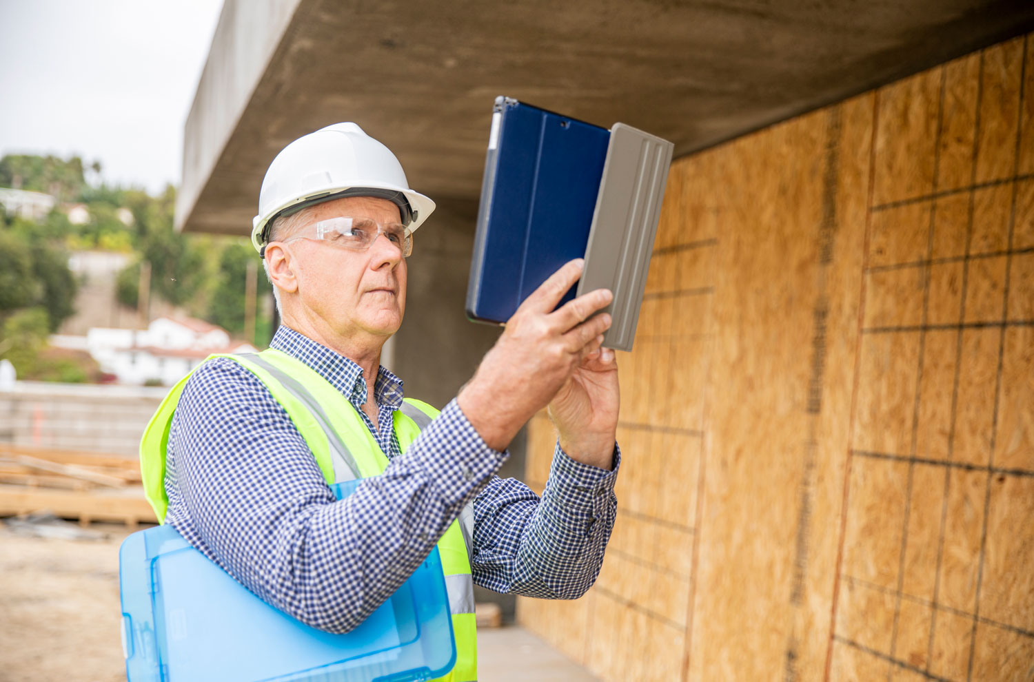 Male home inspector in hard hat using tablet computer by house under construction